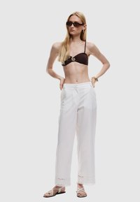 Bruine bikini-top met spiraalaccent, witte wijde broek met kantafwerking en zwarte sandalen met bandjes. Model staat met de handen op de heupen.