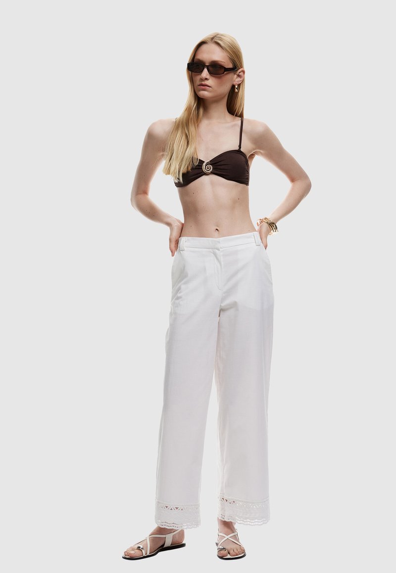 Bruine bikini-top met spiraalaccent, witte wijde broek met kantafwerking en zwarte sandalen met bandjes. Model staat met de handen op de heupen.