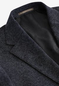 Blazer azul marino oscuro con una tela texturada y tejida, que presenta un cuello con muesca y un forro negro suave. Patrón sutil en el interior.