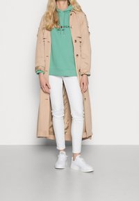 Persona vistiendo un abrigo largo beige sobre una sudadera verde, pantalones ajustados blancos y zapatillas blancas, de pie sobre un suelo gris contra un fondo blanco.