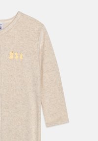 Lys beige baby onesie lavet af blødt stof, med en frontal lynlås, lange ærmer og tre broderede gule ænder på brystet.