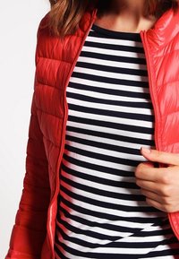 Rote Pufferjacke mit horizontalem Steppdesign über einem navy- und weißgestreiften Oberteil. Die Jacke hat einen hohen Kragen und einen Reißverschluss.