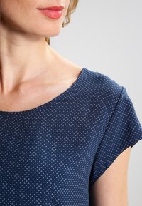 Námorně modrý top s krátkým rukávem a kulatým výstřihem, zdobený texturovaným vzorem malých bílých trojúhelníků po celém povrchu.