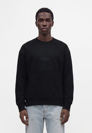 Млад мъж, облечен в черна блуза Emporio Armani и светлосини дънки, стои срещу обикновен светъл фон.