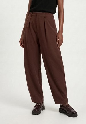 Person som bär höga, löst sittande bruna byxor med veck, svart ärmlös topp och mörkbruna loafers med kraftiga sulor.