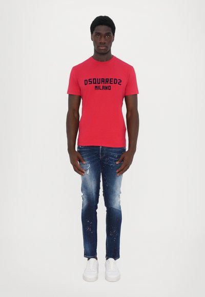 Camiseta de algodón roja con el logotipo en azul "DSQUARED2 MILANO", combinada con jeans azules desgastados con salpicaduras de pintura y zapatillas blancas.
