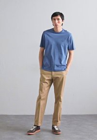 Μπλε βαμβακερό t-shirt με μικρό λογότυπο, καφέ παντελόνι και σκούρα αθλητικά παπούτσια με καφέ λεπτομέρειες. Στάση όρθια μπροστά από ουδέτερο φόντο.