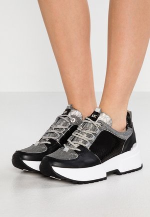 Schwarze und silberne Plateausneaker mit gemusterten Schnürsenkeln, getragen an nackten Füßen vor einfachem hellem Hintergrund.