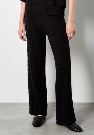 Pantalons larges en tricot noir, à mailles torsadées, avec une texture douce, présentant une taille côtelée. Portés avec des mocassins noirs, sur un fond neutre.
