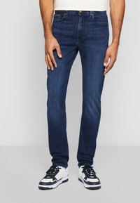 Mies, který má na sobě tmavě modré slim-fit džíny a bílé tenisky s černými akcenty, stojí proti jednoduchému světle šedému pozadí.