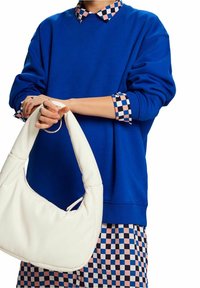 Sudadera azul con collar de cuadros, combinada con un bolso hobo blanco. El bolso tiene una textura suave y forma redondeada, sostenido por el modelo.