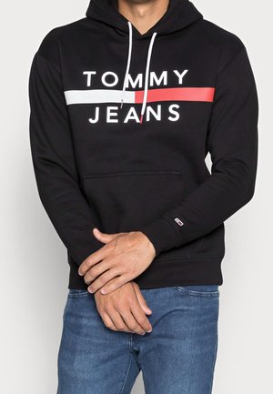 Hombre con sudadera negra Tommy Jeans con una franja horizontal blanca y roja, combinado con vaqueros azules, de pie con los brazos cruzados.