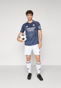 Maglia da calcio blu scuro con il logo "Emirates Fly Better", pantaloni shorts bianchi, scarpini neri, mentre tiene in mano un pallone da calcio a stelle, sfondo minimalista.