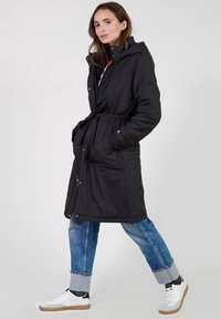 Zwart gewatteerde parka met capuchon, elastische taille en twee voorzakken. Gekozen boven blauwe jeans en witte sneakers, wat een casual stijl toont.