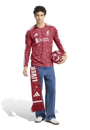 Ung person iført Liverpool FC-langærmet trøje, der holder en tilhørende fodbold og et Liverpool-halstørklæde, stående på en hvid baggrund.