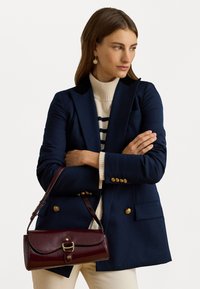 Nő sötétkék blézerben, krémszínű magas nyakú pulóverben és krémszínű nadrágban, sötétvörös bőr kézi táskával arany csattal, az oldalra néz.