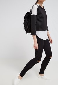 Sudadera en blanco y negro con capas y mangas largas, combinada con jeans skinny negros rasgados y zapatos slip-on blancos. Mochila negra con flecos.