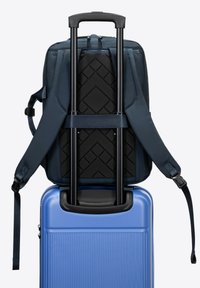 Blauwe harde koffer met een gestructureerd oppervlak. Een marineblauwe rugzak met riemen ligt bovenop, met een zwart bevestigingspaneel voor bagage.