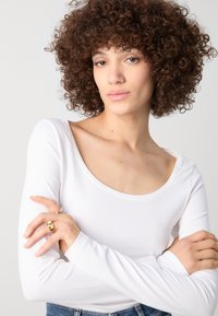 Top blanco de manga larga con escote redondeado, textura suave y ajuste ceñido. El modelo lleva joyas de oro, mostrando anillos en sus dedos.