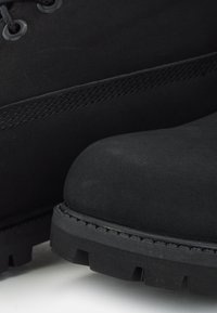 Acercamiento de una bota negra resistente que muestra la parte superior de cuero cosido, suela de goma gruesa con surcos profundos y ojales de metal para los cordones.