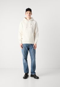 Sudadera blanca con un bolsillo frontal, capucha con cordón y un pequeño logo en el pecho, combinada con jeans azules y zapatos negros.