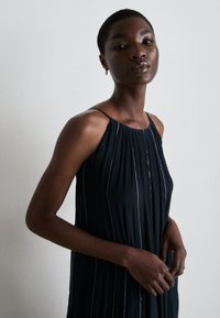 Vestido plissado azul-escuro com alças finas, apresentando riscas verticais brancas. Textura suave e silhueta fluida. Design de decote simples.