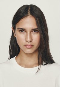 Ritratto in primo piano di una persona con capelli scuri, lunghi e lisci, che indossa una maglietta chiara a girocollo, con un'espressione naturale e una pelle liscia.