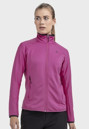 Frau trägt eine pinkfarbene Reißverschlussjacke mit Seitentaschen, steht und schaut seitlich vor einem schlichten Hintergrund.