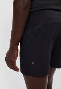 Close-up af en person iført sorte sportsshorts med et lille reflekterende logo nær kanten på venstre lår.