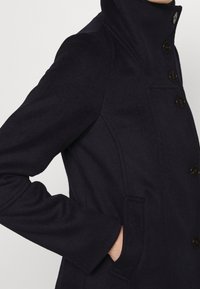 Abrigo de lana marinero con cuello alto, cinco botones y bolsillos laterales. La tela tiene una textura suave con un diseño de corte entallado.