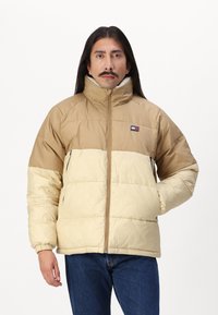 Pufferjack in bruin en crème, heeft een hoge kraag, een rits aan de voorkant en elastische manchetten. Het materiaal lijkt glad met een gestikt ontwerp.