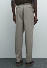 Uomo che indossa pantaloni beige a vestibilità ampia e scarpe nere, in piedi contro una parete grigia chiara e un pavimento scuro macchiato.