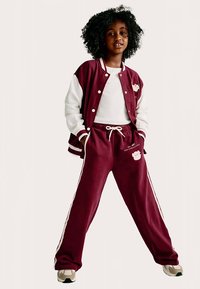 Maroonfarbener College-Blouson mit weißen Ärmeln, kombiniert mit passenden Jogginghosen mit seitlichen Streifen und einem Bärmotiv, beide aus weichem Stoff.