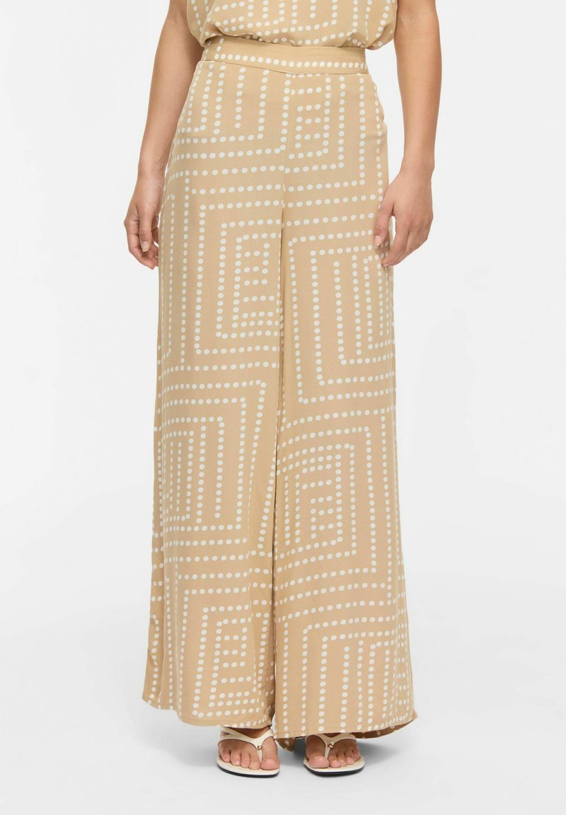 Pantalon beige à jambes larges avec motifs carrés blancs à pois, porté avec des sandales beiges, présenté de la taille aux pieds sur un fond clair.
