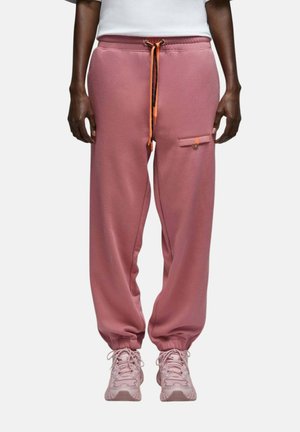 Personne portant un pantalon de survêtement rose avec des poignets élastiques, un cordon orange, une poche latérale avec bouton, et des baskets roses assorties.