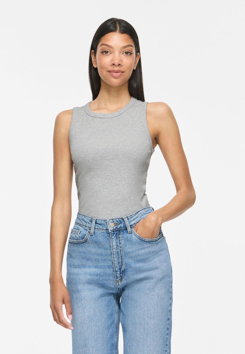 Vrouw met lang donker haar, een grijze mouwloze top en lichtblauwe jeans met hoge taille, staand met één hand in haar zak.