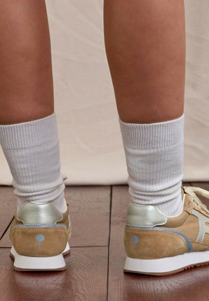 Piernas con calcetines blancos hasta el tobillo y zapatillas de ante beige con detalles plateados, de pie sobre un suelo de madera contra un fondo neutro.