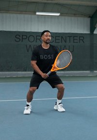 Camiseta deportiva negra con el texto "BOSS", pantalones cortos negros y zapatillas de tenis blancas. Sostenido una raqueta de tenis naranja, posicionado en una cancha.