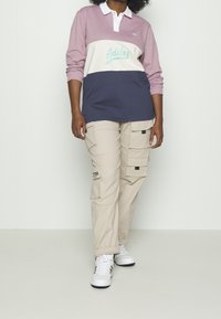 Langärmliges Poloshirt in Pink, Weiß und Marineblau mit "Adidas"-Logo; kombiniert mit beigen Cargo-Hosen mit mehreren Taschen und Reißverschlüssen.