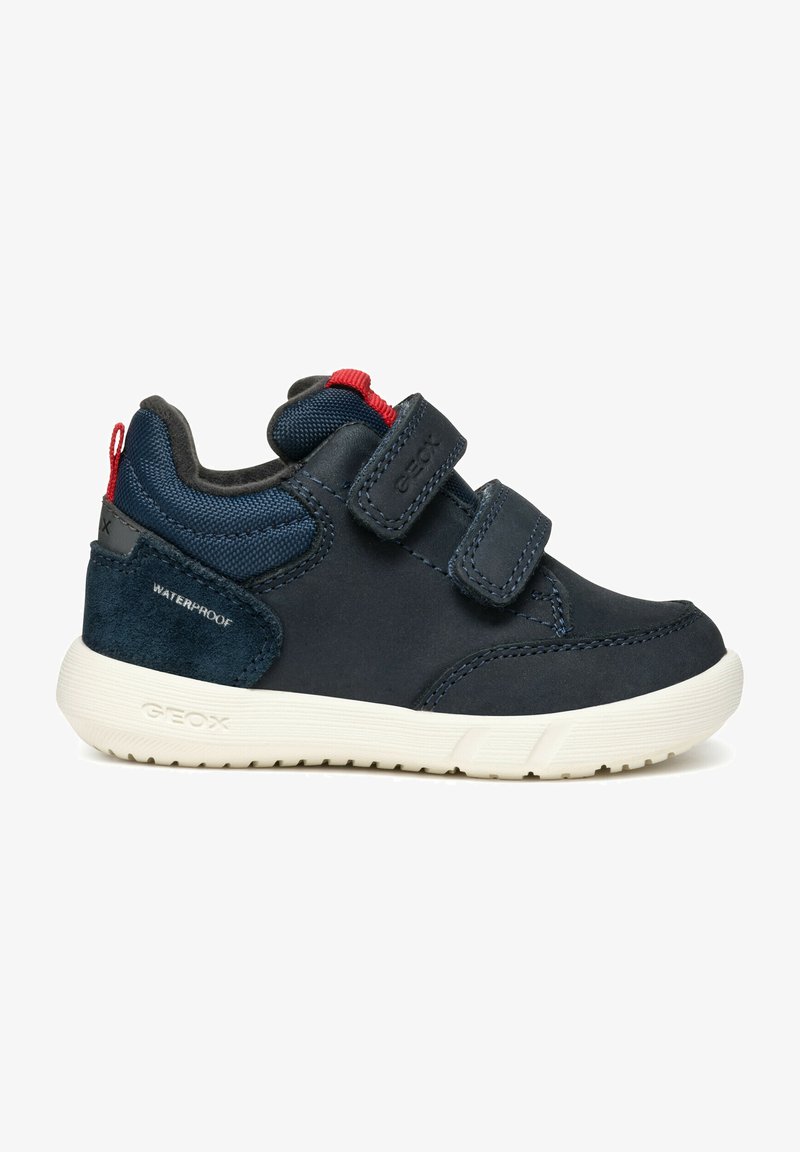 Baskets bleu marine avec des bandes Velcro, en matériau imperméable, accents en daim et semelle en caoutchouc blanche. Présente un onglet rouge à l'arrière.