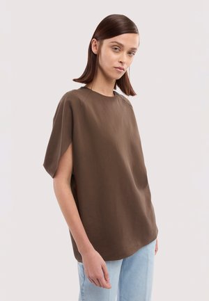 Frau mit glatten braunen Haaren, die ein lockeres braunes Kurzarmshirt und hellblaue Jeans trägt, steht vor einem schlichten hellen Hintergrund.