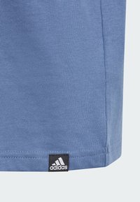 Camiseta de algodón azul con textura acanalada, dobladillo recto y una etiqueta del logo de Adidas en negro en la esquina inferior izquierda.