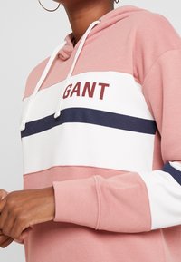 Haut à capuche rose avec des rayures blanches et bleu marine, arborant le texte "GANT" en bordeaux. Fabriqué en tissu doux, il comprend une capuche avec cordon et des poignets côtelés.
