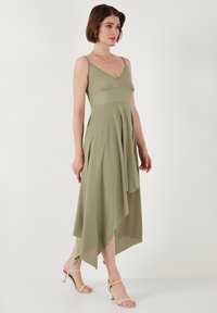 Asymmetrical olive green dress with thin straps, textured fabric, and a flowy silhouette; paired with beige sandals.