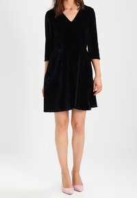 Vestido de terciopelo negro con mangas tres cuartos, escote en V, falda acampanada y bolsillos laterales. Llevado con tacones rosas.