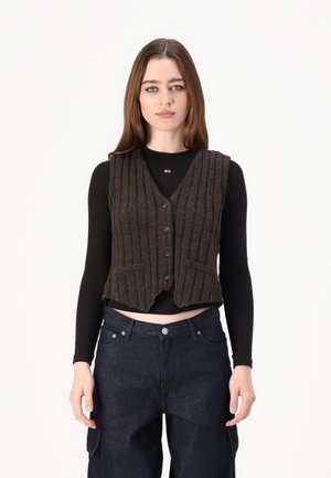 Brun ribbet vest med V-hals, knappelukking og lommer, båret over en svart langermet topp og blå denimjeans.