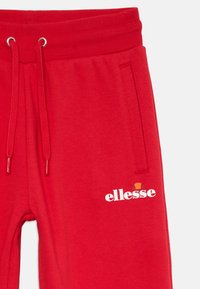 Červené tepláky s elastickým pásom, sťahovacími šnúrkami, bočnou vreckom a bielym logom "ellesse" s oranžovou a červenou grafikou nad ním.