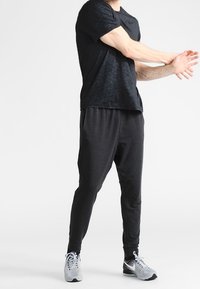 Czarna sportowa koszulka z teksturowanym wykończeniem, w zestawie z czarnymi spodniami dresowymi. Zauważalne szare buty do biegania z siateczkowym wzorem i sznurowadłami.