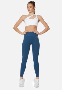 Bra sportivo bianco asimmetrico con ritaglio; leggings alti blu scuro; texture liscia; indossato con sneakers bianche.