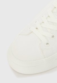 White canvas sneaker featuring a round toe, flat laces, smooth rubber sole, and subtle stitching details near the eyelets.